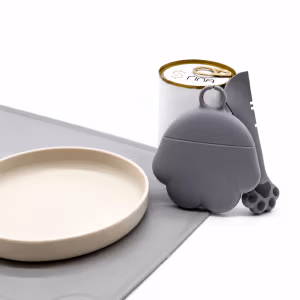 Beiger Katzennapf auf grauer Unterlage neben einer Rina-Futterdose, einem grauen Silikondeckel und einem Löffel mit Pfotenmotiv.